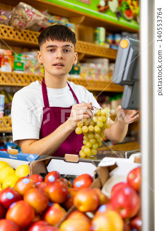 Young male supermarket worker weighing on the scales green grapes in grocery shop Young male supermarket worker weighing on the scales green grapes in grocery shop 140553764