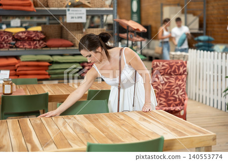 Pretty brunette girl in white dress chooses wooden table for country house in garden store Pretty brunette girl in white dress chooses wooden table for country house in garden store 140553774