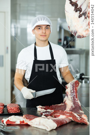 Guy employee of butcher shop cuts meat carcass into small pieces of meat for retail sale in store Guy employee of butcher shop cuts meat carcass into small pieces of meat for retail sale in store 140554042