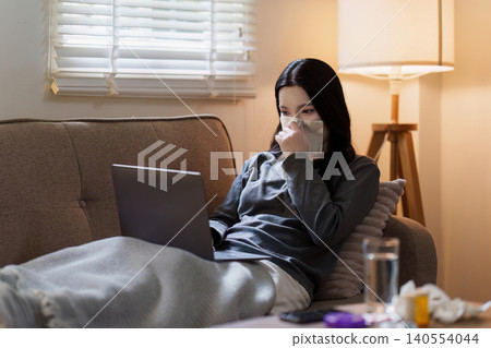 Asian woman working on laptop while sick with cold on couch at home Asian woman working on laptop while sick with cold on couch at home 140554044