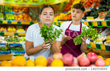 Doubted young female customer asking advice from male market assistant while buying greens in supermarket Doubted young female customer asking advice from male market assistant while buying greens in supermarket 140554045