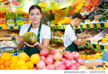 Positive young saleswoman holding fresh melon during offering fresh fruits Positive young saleswoman holding fresh melon during offering fresh fruits 140554195