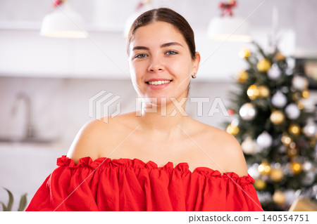Portrait of happy young female in red dress smiling at camera in front of decorated Christmas tree at home Portrait of happy young female in red dress smiling at camera in front of decorated Christmas tree at home 140554751