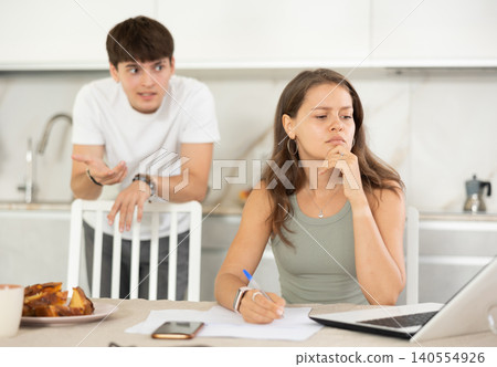 Young girl sitting at the table while her boyfriend is quarreling with her standing behind Young girl sitting at the table while her boyfriend is quarreling with her standing behind 140554926