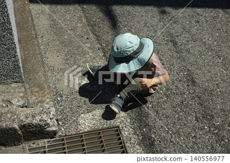 A child wearing a hat sits by the roadside playing with sand and stones. A child wearing a hat sits by the roadside playing with sand and stones. 140556977