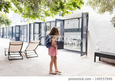 Girl child standing on tiled courtyard, carrying navy blue backpack toward glass doors, copy space Girl child standing on tiled courtyard, carrying navy blue backpack toward glass doors, copy space 140557940