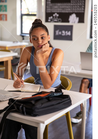 Teenage girl sitting at desk in classroom, holding pencil over notebook near laptop Teenage girl sitting at desk in classroom, holding pencil over notebook near laptop 140558203