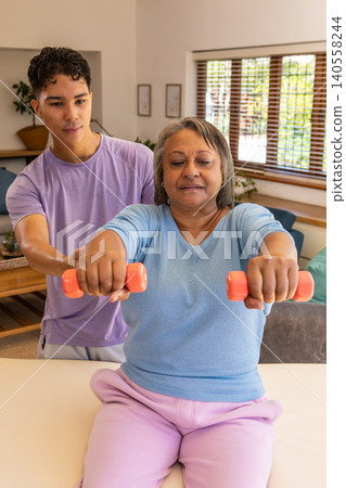 Hispanic trainer guiding senior female client lifting orange dumbbells on therapy table at home Hispanic trainer guiding senior female client lifting orange dumbbells on therapy table at home 140558244