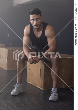 Muscular man sitting on wooden plyometric box on rubber mat floor in dim gym wearing smartwatch Muscular man sitting on wooden plyometric box on rubber mat floor in dim gym wearing smartwatch 140558294