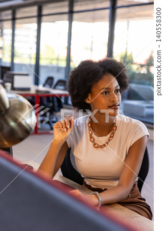 African american woman sitting at office desk holding pencil gazing at monitor near metallic lamp African american woman sitting at office desk holding pencil gazing at monitor near metallic lamp 140558300