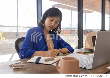 Asian woman writing in notebook with metallic pen at modern office with laptop fabric swatches Asian woman writing in notebook with metallic pen at modern office with laptop fabric swatches 140558304