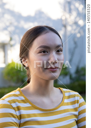 Asian woman standing in residential garden wearing striped shirt and stud earrings Asian woman standing in residential garden wearing striped shirt and stud earrings 140558320