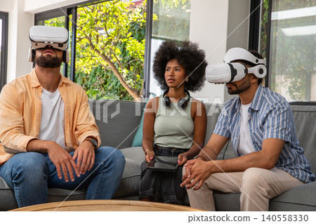 Diverse friends sitting on sofa in living room wearing vr headsets and using tablet Diverse friends sitting on sofa in living room wearing vr headsets and using tablet 140558330