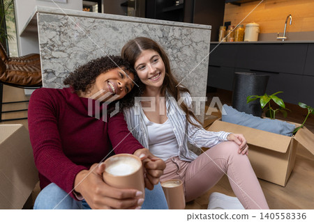 Diverse female friends sitting by kitchen island holding ceramic mugs among moving boxes, plant Diverse female friends sitting by kitchen island holding ceramic mugs among moving boxes, plant 140558336