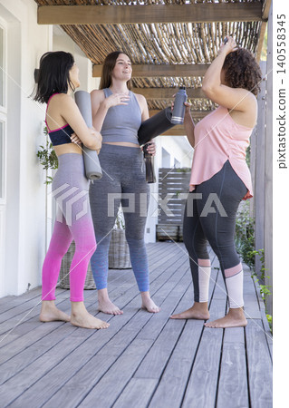 Diverse female friends holding yoga mats and water bottles chatting under bamboo pergola on porch Diverse female friends holding yoga mats and water bottles chatting under bamboo pergola on porch 140558345