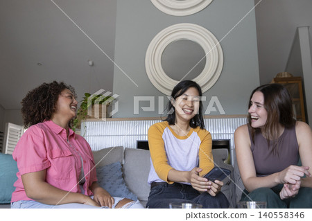 Diverse female friends chatting on couch in modern living room holding smartphone near glass cups Diverse female friends chatting on couch in modern living room holding smartphone near glass cups 140558346