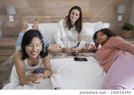 Diverse female friends relaxing on bed in bedroom with wood-paneled wall holding smartphones 140558349