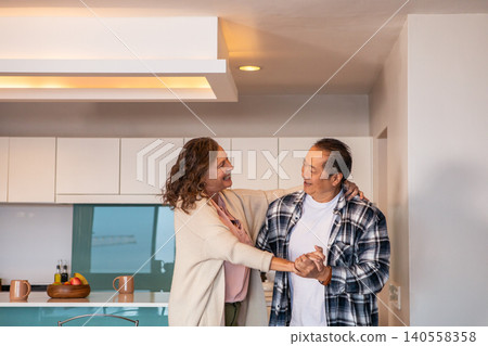 Diverse senior couple dancing holding hands in kitchen near countertop with fruit bowl Diverse senior couple dancing holding hands in kitchen near countertop with fruit bowl 140558358