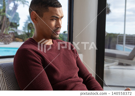 Mid adult African American man wearing sweater gazing toward outdoor pool via glass door at home 140558372