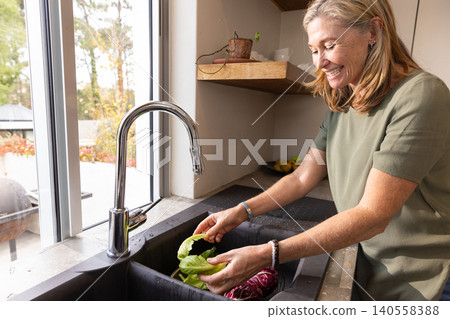Senior woman washing lettuce and radicchio under chrome faucet at sink by window Senior woman washing lettuce and radicchio under chrome faucet at sink by window 140558388