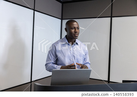 African american man gesturing behind lectern using laptop in office featuring frosted glass African american man gesturing behind lectern using laptop in office featuring frosted glass 140558393