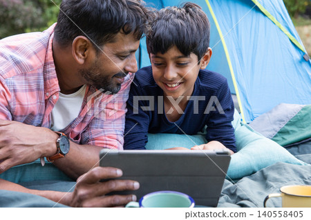 Indian father with son watching tablet while lying on camping mats at entrance of blue tent Indian father with son watching tablet while lying on camping mats at entrance of blue tent 140558405