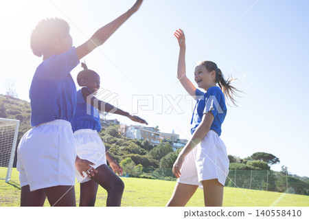 Diverse teenage teammates celebrating on soccer field by goal wearing blue jerseys and shorts Diverse teenage teammates celebrating on soccer field by goal wearing blue jerseys and shorts 140558410