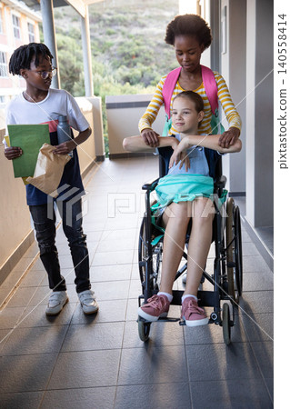 Diverse classmates pushing wheelchair walking down corridor holding folder bag water bottle 140558414
