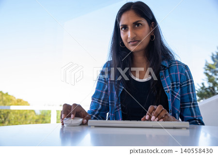 Indian woman typing on white keyboard and using wireless mouse at balcony desk Indian woman typing on white keyboard and using wireless mouse at balcony desk 140558430