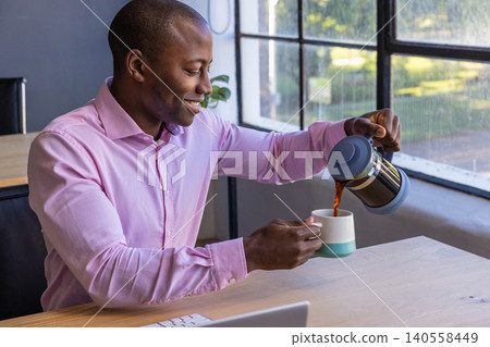 African american man pouring coffee from french press at desk near window, copy space African american man pouring coffee from french press at desk near window, copy space 140558449