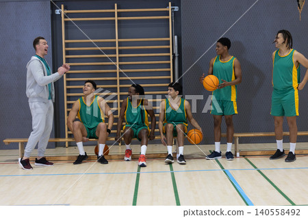 Diverse male basketball team listening to coach shouting in gym with basketballs and bench Diverse male basketball team listening to coach shouting in gym with basketballs and bench 140558492
