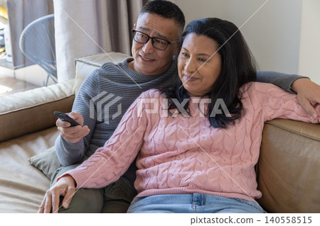 Senior couple sitting on leather couch in living room with tv remote with throw pillow Senior couple sitting on leather couch in living room with tv remote with throw pillow 140558515