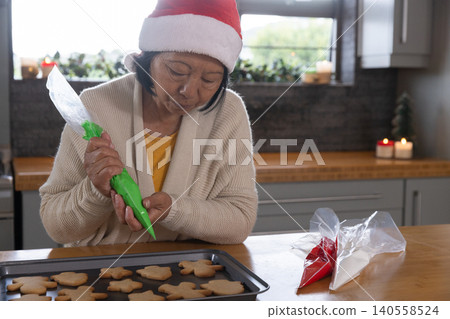 Senior Asian woman wearing Santa hat decorating cookies using green icing bag at kitchen counter Senior Asian woman wearing Santa hat decorating cookies using green icing bag at kitchen counter 140558524