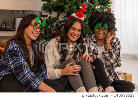Diverse female friends sitting on wooden floor by christmas tree in glasses and santa headband Diverse female friends sitting on wooden floor by christmas tree in glasses and santa headband 140558528