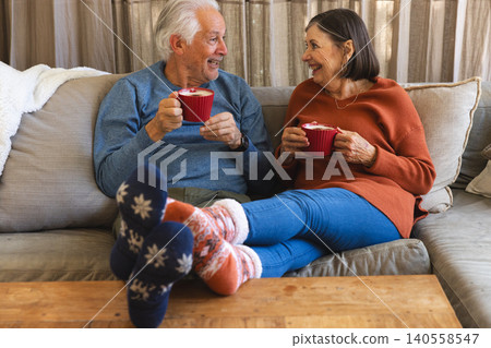 Senior couple sitting on beige cushioned sofa in cozy living room holding red ribbed mugs 140558547