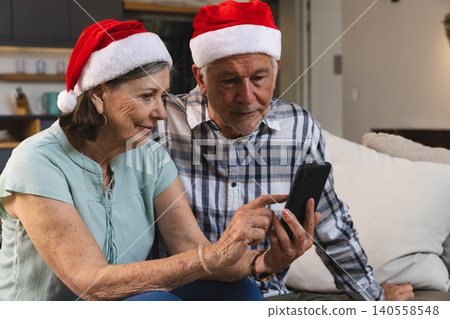 Senior couple wearing Santa hats scrolling smartphone on sofa with white pillow near kitchen mugs Senior couple wearing Santa hats scrolling smartphone on sofa with white pillow near kitchen mugs 140558548