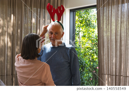 Couple adjusting red reindeer antler headband near living room window and beige curtains Couple adjusting red reindeer antler headband near living room window and beige curtains 140558549