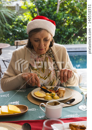 Senior woman wearing Santa hat cutting holiday meal at patio beside pool with plate of food Senior woman wearing Santa hat cutting holiday meal at patio beside pool with plate of food 140558551