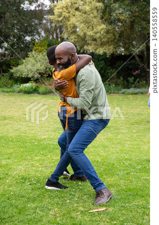 African american father and child lifting and hugging on lawn in park, green trees backdrop 140558559