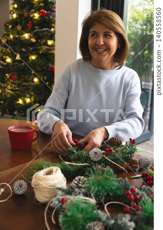 Senior woman sitting at table in living room making holiday wreath with pinecones and red mug 140558560