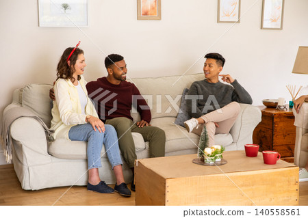Diverse friends sitting on sofa in living room, chatting around holiday centerpiece with mugs Diverse friends sitting on sofa in living room, chatting around holiday centerpiece with mugs 140558561