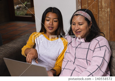 Diverse mother and daughter researching recipes on laptop while sitting on living room couch Diverse mother and daughter researching recipes on laptop while sitting on living room couch 140558572