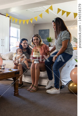 Diverse female friends with baby marking thirtieth birthday in living room with cake, balloons Diverse female friends with baby marking thirtieth birthday in living room with cake, balloons 140558579