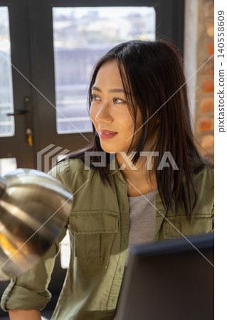 Asian woman looking thoughtfully to left sitting at desk in modern workspace near laptop and lamp Asian woman looking thoughtfully to left sitting at desk in modern workspace near laptop and lamp 140558609