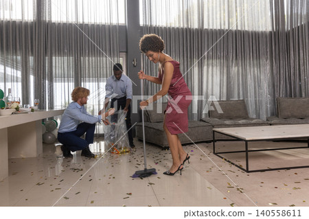 Diverse friends tidying living room with broom picking confetti into trash bag, copy space Diverse friends tidying living room with broom picking confetti into trash bag, copy space 140558611