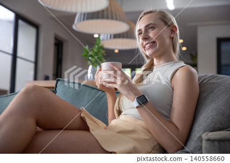 Woman holding mug on gray sofa in living room wearing smartwatch under pendant lamps Woman holding mug on gray sofa in living room wearing smartwatch under pendant lamps 140558660