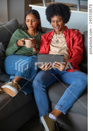 Indian and african american couple sitting on gray sofa in living room with tablet and mug 140558678