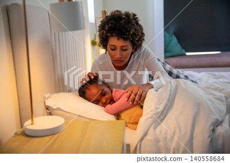 Diverse mother and daughter kneeling on bed, comforting child under duvet with teddy bear Diverse mother and daughter kneeling on bed, comforting child under duvet with teddy bear 140558684