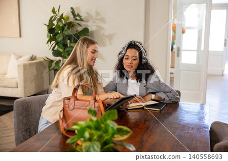 Diverse female colleagues pointing at tablet, notebook in home workspace Diverse female colleagues pointing at tablet, notebook in home workspace 140558693