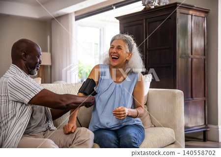 Diverse senior couple using blood pressure cuff on beige sofa in living room beside lamp, armoire Diverse senior couple using blood pressure cuff on beige sofa in living room beside lamp, armoire 140558702